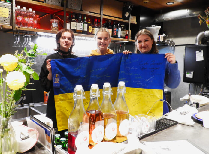 メッセージが書かれた国旗を持つウクライナ料理店「バブーシャ　レイ」のスタッフ。左からミヤさん、ヴィクトリヤさん、ジェーニャさん＝東京都武蔵野市（辻本奈緒子撮影）